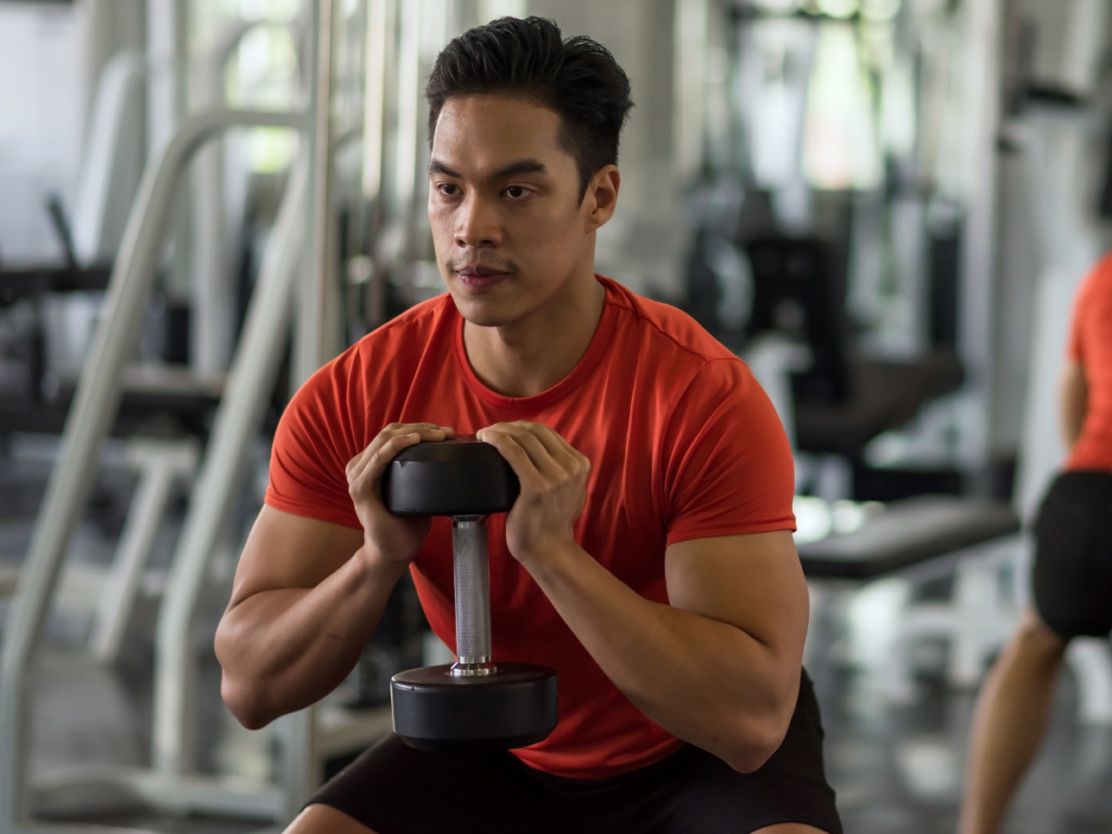 Photo of middle-aged man in a red shirt participating in a group exercise class using a dumbbell mid-squat.