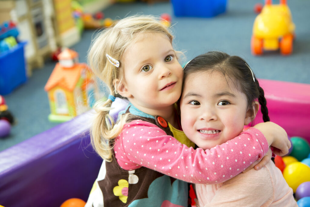 Blonde toddler wraps her arms around another toddler.
