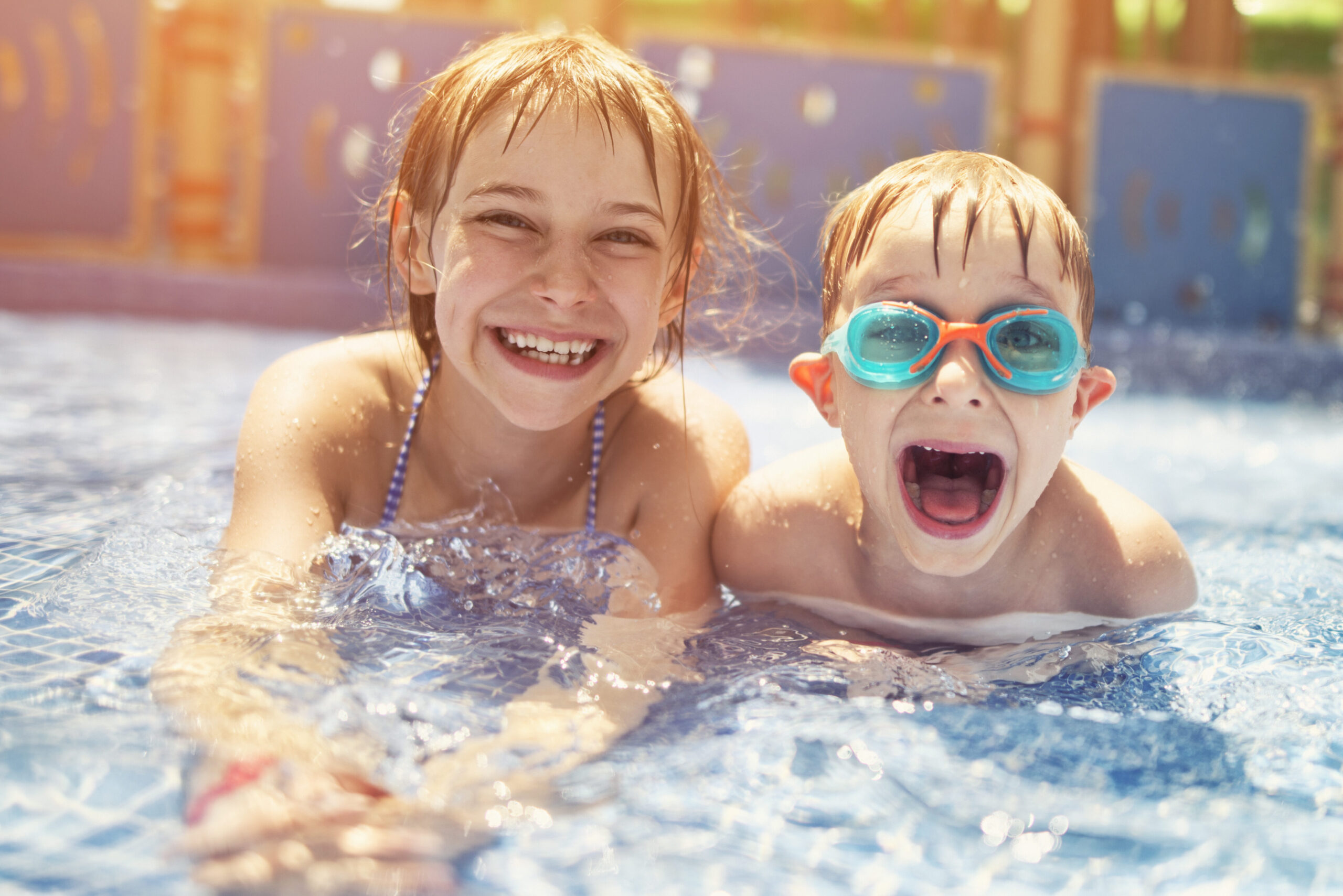 Little girl and her brother lying on front in shallow water in water park. The kids are laughing at camera, the boy is wearing swimming goggles.